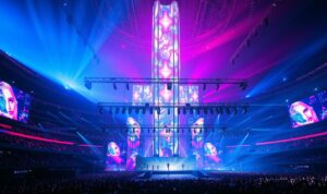 An extravagant stage design with a towering holographic structure at the center, casting vibrant hues of blue, violet, and pink across the arena. Sleek, futuristic screens project dazzling visuals, complementing the dynamic dance troupe's intricate choreography. In the foreground, a mesmerizing array of lighting fixtures and rigging create an electrifying atmosphere, while the audience is enveloped in a captivating sonic experience. The scene exudes a sense of technological innovation and artistic splendor, reflecting the cutting-edge production of Eras Tour, Taylor Swift's highly anticipated world tour.